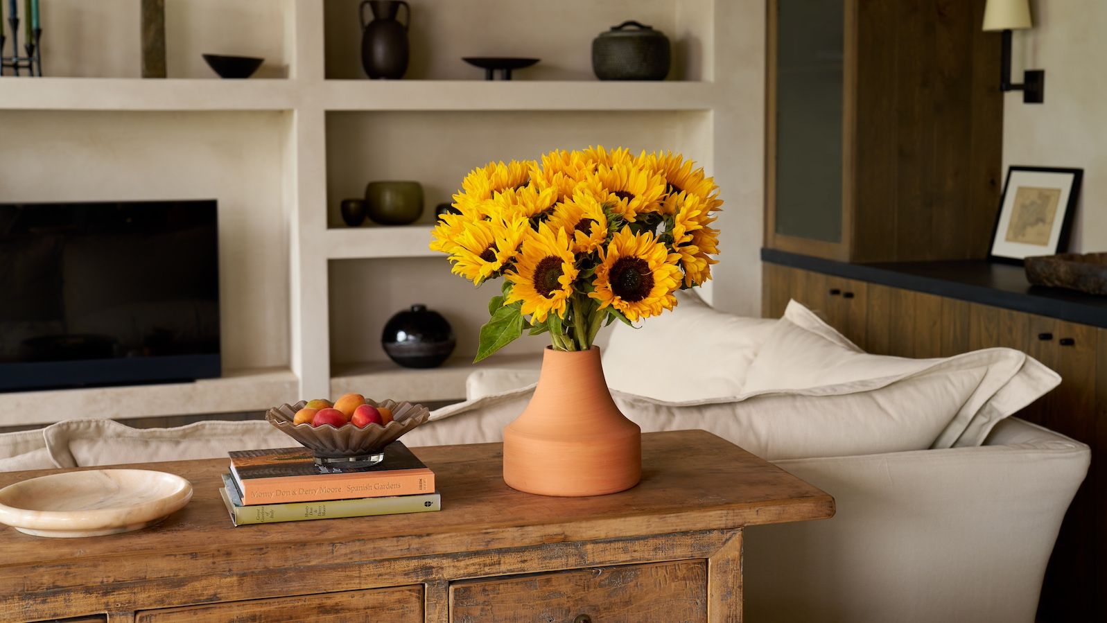 A bouquet of sunflowers in a terracotta vase sit on a sideboard in a minimalist room
