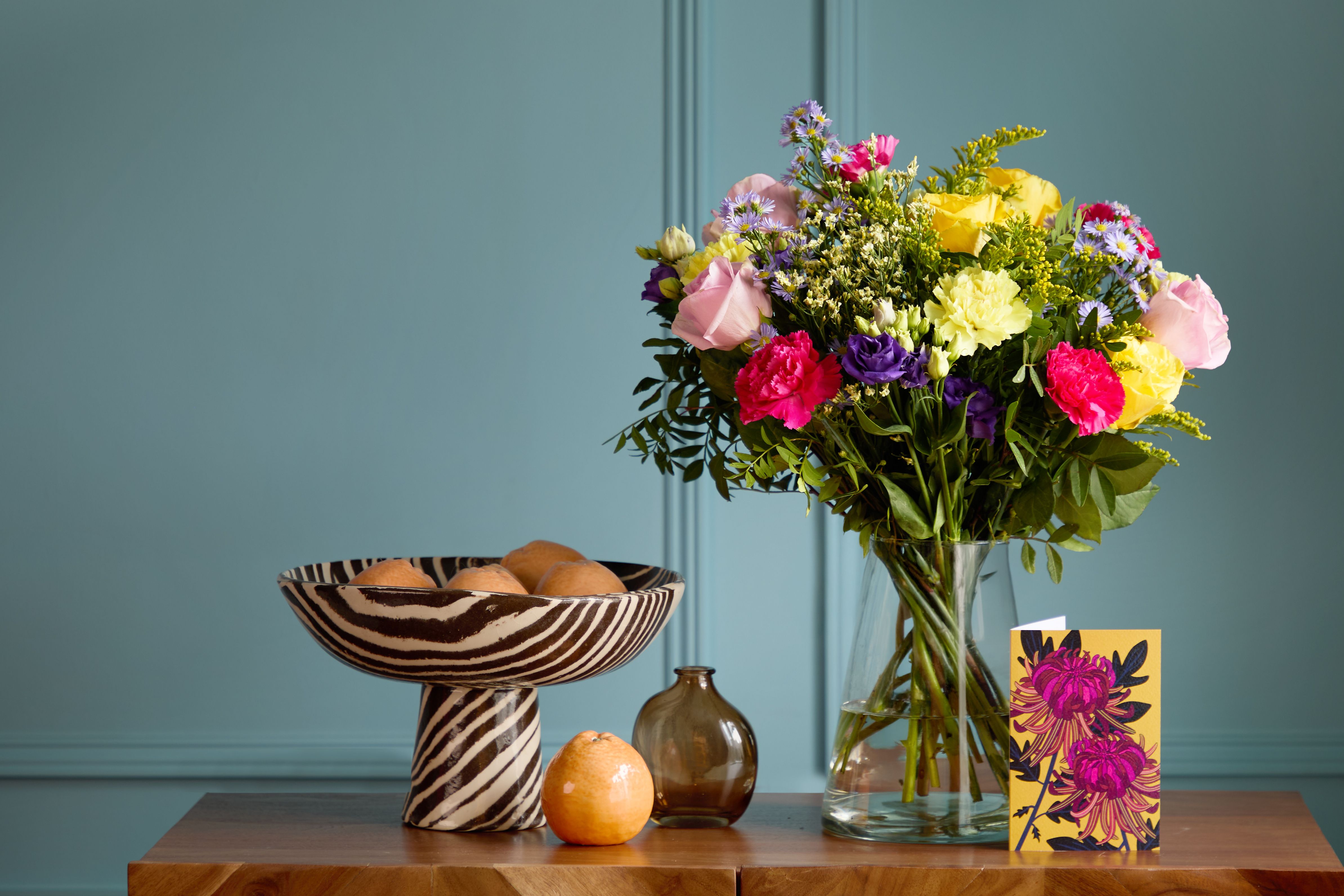 Arena Flowers' 'Dancing 'Queen bouquet, a mix of yellow, red and purple flowers, in a vase against blue wall