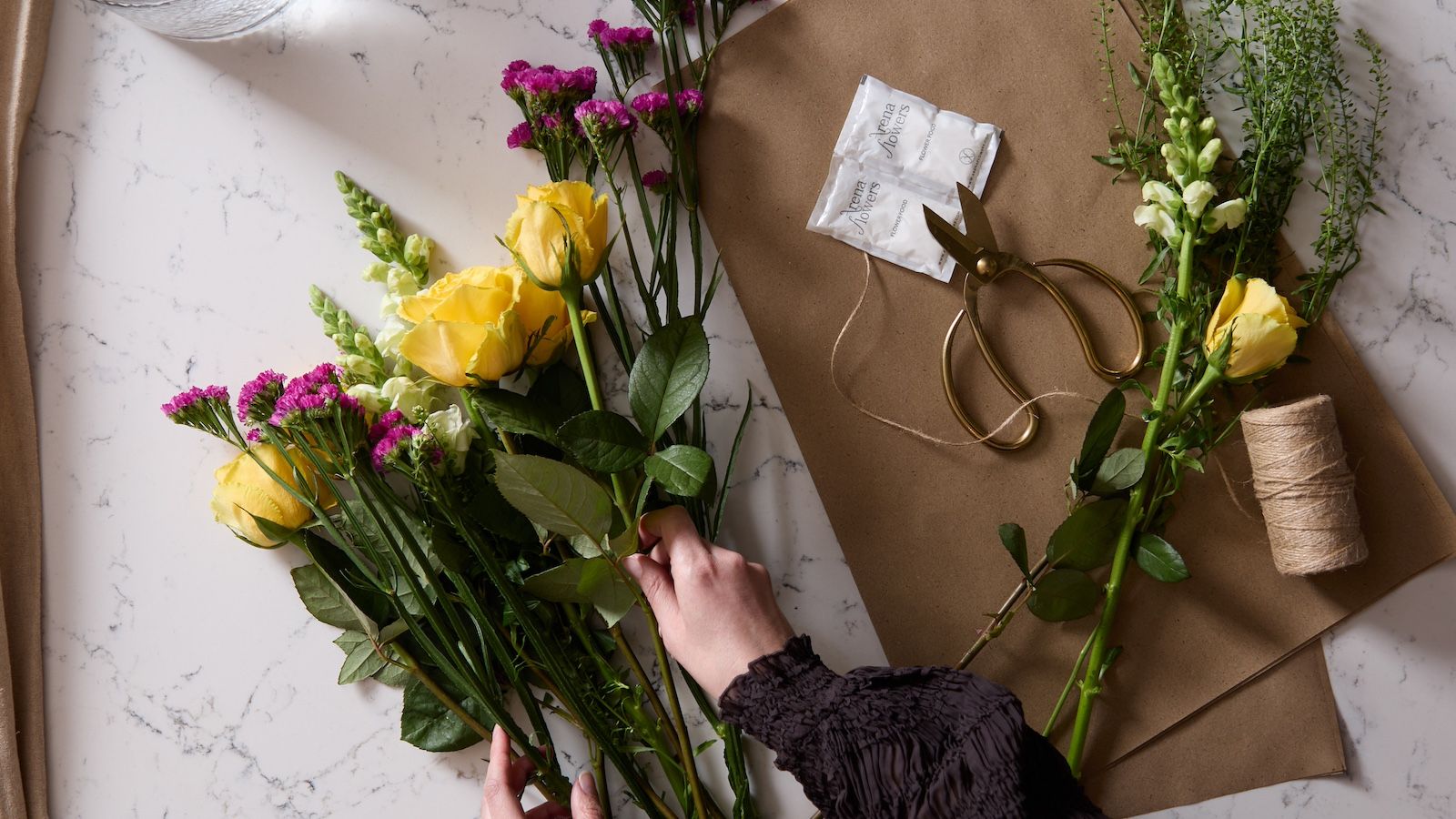 An aeriel shot of a bouquet being arranged, with scissors, flower food, string and stems arranged on a marble worktop