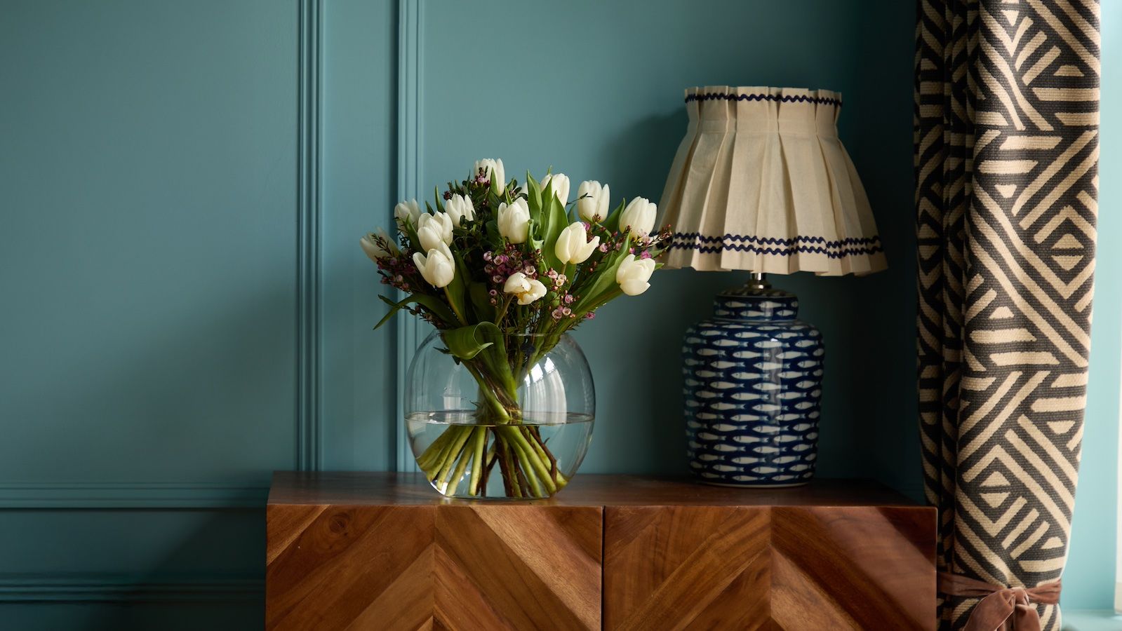 A vase of white tulips and pink waxflowers sits on a sideboard next to a blue patterned lamp