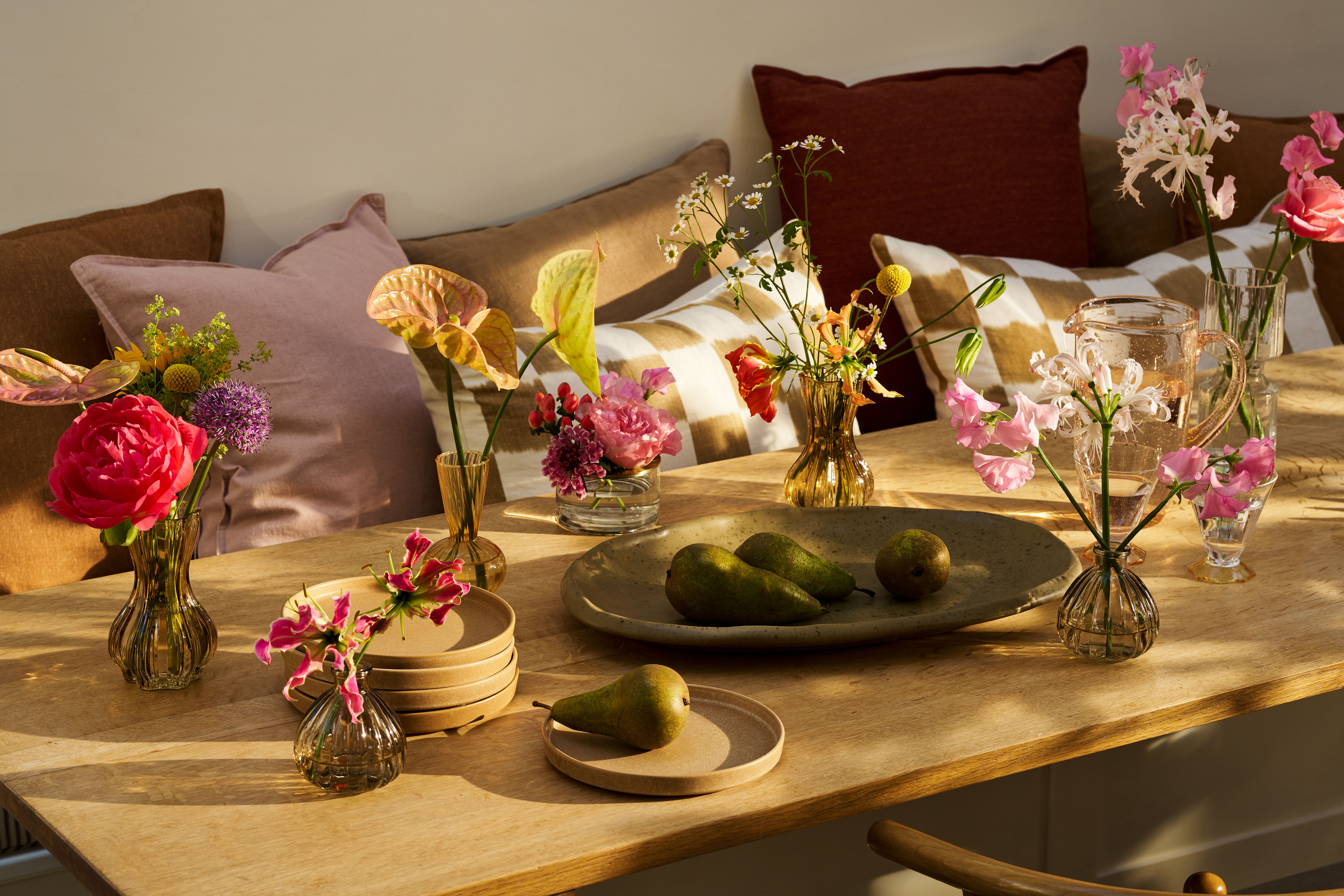 A series of flowers in bud vases on a dining table, with plates and a bowl of pears